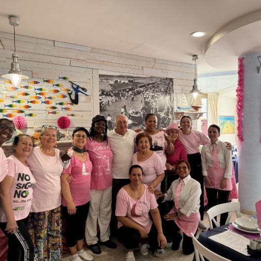 Gruppo di persone in rosa, sorridenti in un ristorante decorato con pesci colorati.