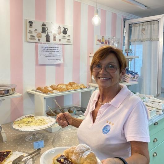 Donna sorridente serve dolci in una pasticceria con sfondo a righe rosa.