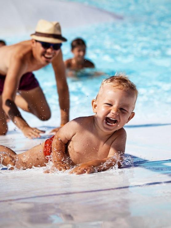 Bambino felice gioca in piscina, seguito da un adulto sorridente.